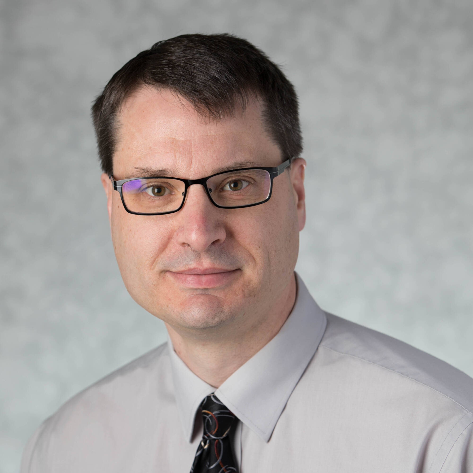 Professional headshot of William wearing glasses, a light gray shirt, and a patterned tie, facing the camera with a neutral expression against a gray background